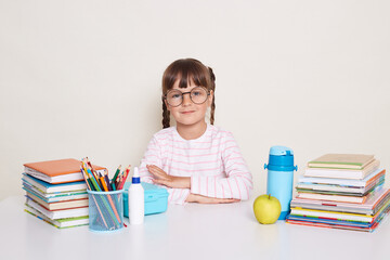 Portrait of serious little schoolgirl with pigtails and glasses wearing striped shirt sitting at the desk surrounded with books and pens, keeps hands folded, looking at camera, waiting for teacher.