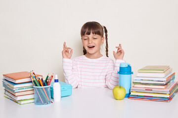 Happy satisfied hopeful little schoolgirl with pigtails wearing white shirt sitting at the desk with closed eyes, crossed fingers, making desire, posing surrounded with books and other school supplies