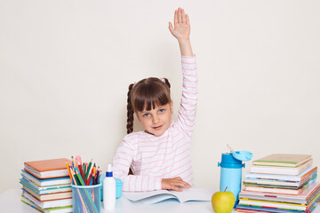 Horizontal shot of excellent little schoolgirl with dark hair and braids sitting at table with raised arm, answering, child knows right answer, looking at camera, being surrounded with books.
