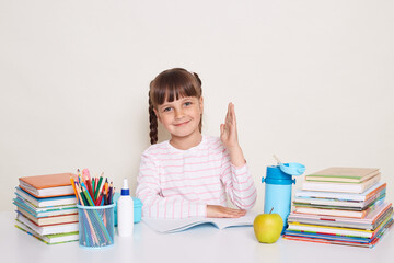 Indoor shot of smiling delighted little schoolgirl with dark hair and braids sitting at table surrounded with books and raised arm, child want to answer during lesson.