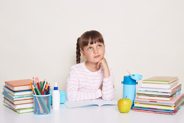 Thoughtful little schoolgirl wearing striped shirt sitting at the desk surrounded with books and other school supplies, looking away, thinking during lesson or while dong homework, holding chin.
