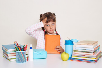 Indoor shot of sad sick little schoolgirl with dark hair and braids, little girl touching her forehead, feels unwell, suffering headache, sitting at table surrounded with books.