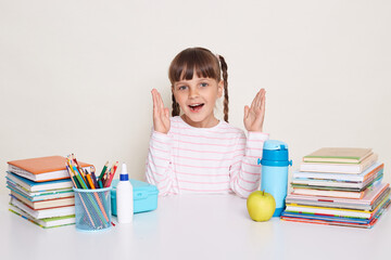 Indoor shot of happy optimistic little schoolgirl with pigtails wearing white shirt sitting at the desk with raised arms, showing big size, surrounded with books and other school supplies