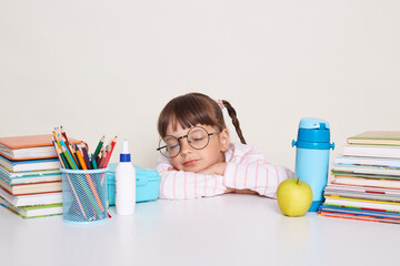 Portrait of sleepy little schoolgirl with pigtails wearing striped shirt sitting at the desk with closed eyes, sleeping during break, being surrounded with books and other school supplies.