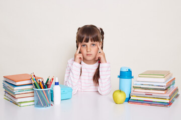 Image of sad serious little schoolgirl with pigtails wearing striped shirt, keeps fingers on temples, looking at camera, sitting at the desk surrounded with books and other school supplies.