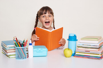 Photo of excited amazed little schoolgirl with dark hair and braids sitting at table surrounded with books and holding book and looking at camera with open mouth and expressing excitement.