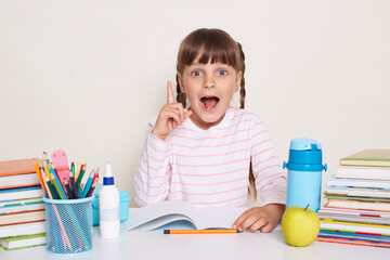 Indoor shot of excited little schoolgirl with dark hair and braids sitting at table with raised finger, pointing up, having excellent idea, screaming eureka, posing isolated over white background.