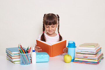 Horizontal shot of smiling pretty little schoolgirl with pigtails wearing striped shirt sitting at the desk surrounded with books and other school supplies and reading during literature lesson.