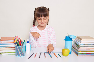 Indoor shot of smiling little schoolgirl with dark hair and braids sitting at table surrounded with books, having mathematics lesson, counting colored pencils, studying.