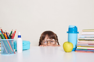 Funny portrait of little schoolgirl with pigtails wearing striped shirt sitting hiding from desk, spying , having fun during break, female child in glasses looking at camera.