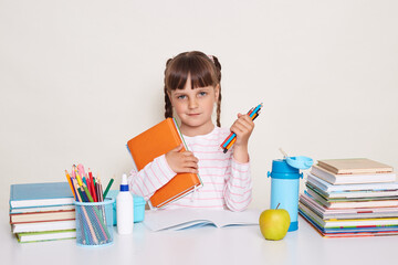 Portrait of cute charming little schoolgirl with dark hair and braids sitting at table, holding book and pencils, looking at camera, waiting her teacher and starting lesson.