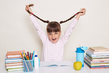 Photo of happy positive little schoolgirl with dark hair and braids sitting at table surrounded with books and playing, having fun during break, raised her pigtails and screaming, looking at camera.