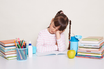 Horizontal shot of sleepy exhausted little schoolgirl with dark hair and braids sitting at table,...