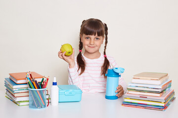 Horizontal shot of smiling optimistic little schoolgirl with pigtails wearing striped shirt sitting at the desk surrounded with books and other school supplies, holding fresh fruit, looking at camera.