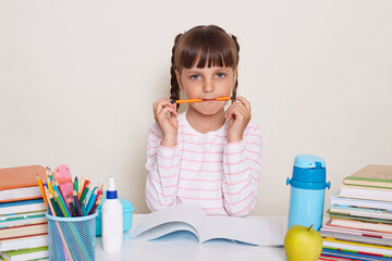Portrait of funny little schoolgirl with pigtails wearing striped shirt sitting at the desk surrounded with school supplies, writing in exercises, holding pen in mouth.