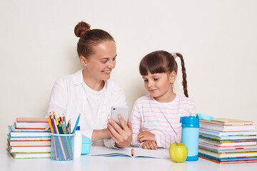 Horizontal shot of Caucasian woman with her daughter or pupil sitting at table with books doing homework, expressing positive emotions, smiling while studying, using mobile phone for doing tasks.