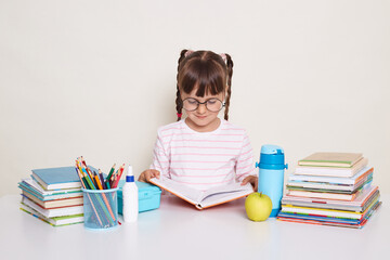 Image of serious attentive little schoolgirl with dark hair and braids sitting at table surrounded with books and reading, doing tasks from literature during lesson.