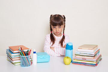 Horizontal shot of sad little schoolgirl with dark hair and braids sitting at table surrounded with books, looking at camera with pout lips and upset eyes, expressing sadness and sorrow.