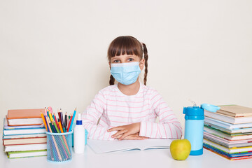 Horizontal shot of little schoolgirl with dark hair and braids sitting at table surrounded with books, waiting for lesson start, looking at camera, having flu symptoms, wearing protective mask.