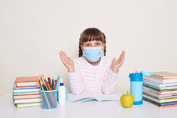 Portrait of little schoolgirl with pigtails wearing protective mask sitting at the desk surrounded with books, spread arms, visiting school during flu virus epidemic.