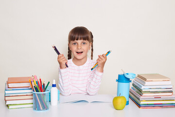 Photo of amazed little schoolgirl with pigtails wearing striped shirt sitting at the desk surrounded with school supplies, holding pens or pencils, looking at camera with optimistic happy expression.