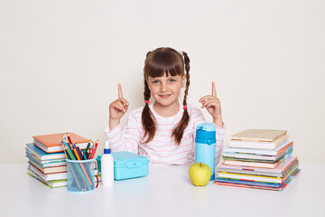 Image of smiling positive optimistic little schoolgirl with dark hair and braids sitting at table surrounded with books, pointing fingers up, looking at camera, posing isolated over white background.