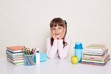 Portrait of smiling pretty cute little schoolgirl with dark hair and braids sitting at table surrounded with books, looking at camera and keeps palms on cheeks, being in good mood.