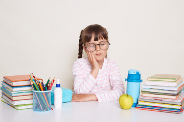 Horizontal shot of sleepy little schoolgirl with dark hair and braids sitting at desk surrounded with books isolated over white background, keeps eyes closed.