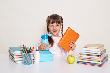 Horizontal shot of winsome positive little schoolgirl with dark hair and braids, holding book and blue water bottle, sitting at table surrounded with books against white wall.