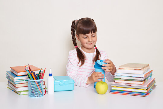 Image of smiling happy little schoolgirl with dark hair and braids sitting at table surrounded with books, holding bottle with water, posing during break isolated over white background.