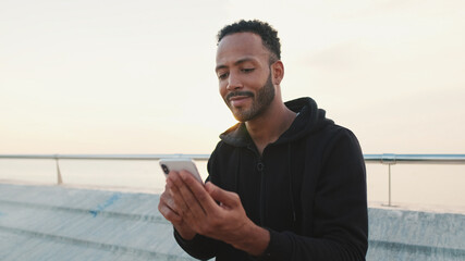 Close up, young smiling bearded male athlete dressed in hoodie uses cellphone after training while sitting on the embankment