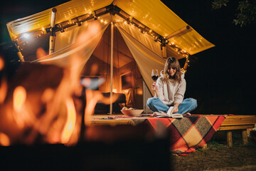 Fototapeta na wymiar Smiling Woman freelancer drinking wine and read book sitting in cozy glamping tent in autumn evening. Luxury camping tent for outdoor holiday and vacation. Lifestyle concept