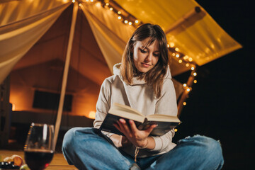 Smiling Woman freelancer drinking wine and read book sitting in cozy glamping tent in autumn...