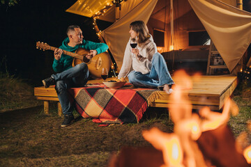 Obraz na płótnie Canvas Happy couple relaxing in glamping on autumn evening, drinking wine and playing guitar near cozy bonfire. Luxury camping tent for outdoor recreation and recreation. Lifestyle concept