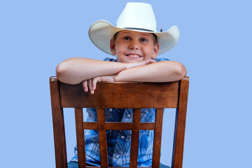 Young child cowboy wears blue plaid shirt and white cowboy hats sits on chair with arms crossed,...