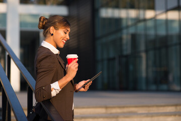 Young smiling businesswoman drinking coffee and using tablet while standing on the stairs in front of corporate office building