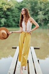 A young woman in a hippie look and eco-dress dancing outdoors by the lake wearing a hat and yellow pants in the summer sunset