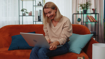 Obraz premium Overjoyed caucasian girl at home sitting on couch with laptop scream in delight raise hands in triumph winner gesture celebrate success win money in lottery. Excited young woman get online good news