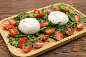 Fresh Italian buratta cheese served with arugula salad and cherry tomatoes on a wooden plate stands on the table