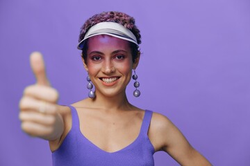 Portrait of a sporty fashion woman posing smiling with teeth and pointing a finger at the camera in a purple yoga tracksuit and a transparent cap on a purple monochrome background