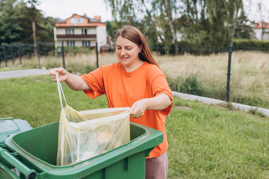 Young happy woman putting garbage in green trash bin on back yard background. Smiling women holding yellow plastic trash bag. Environmental conservation saving planet from contamination