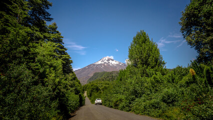 camioneta en un camino rural rodeado de arboles verdes. al fondo se ve una montaña o volcán...