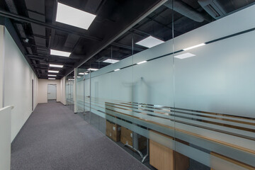 A wide corridor of a modern office with furniture behind glass partitions.