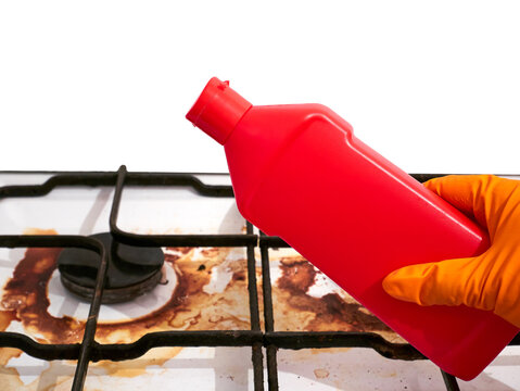 A hand in a rubber glove holds red bottle with chemical clean on the background of a dirty gas stove. The detergent in the bottle. Washing from the dirt of the kitchen gas stove.