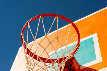 Colourful orange basketball backboard with red hoop and a net against blue sky low angle view.