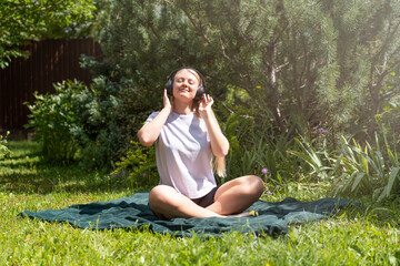 A young blonde girl is sitting on the grass and listening to music in wireless headphones