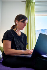 Vertical shot of Caucasian woman with focused face looking at computer from bedroom