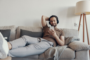 Handsome young man in headphones using smart phone while relaxing on the couch at home