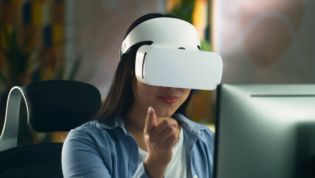 Woman in VR headset sitting at the table at home and working remotely on computer or surfing the internet in spare time. Virtual reality.