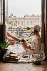 Beautiful young woman in bathrobe pouring some champagne while relaxing on the balcony - obrazy, fototapety, plakaty